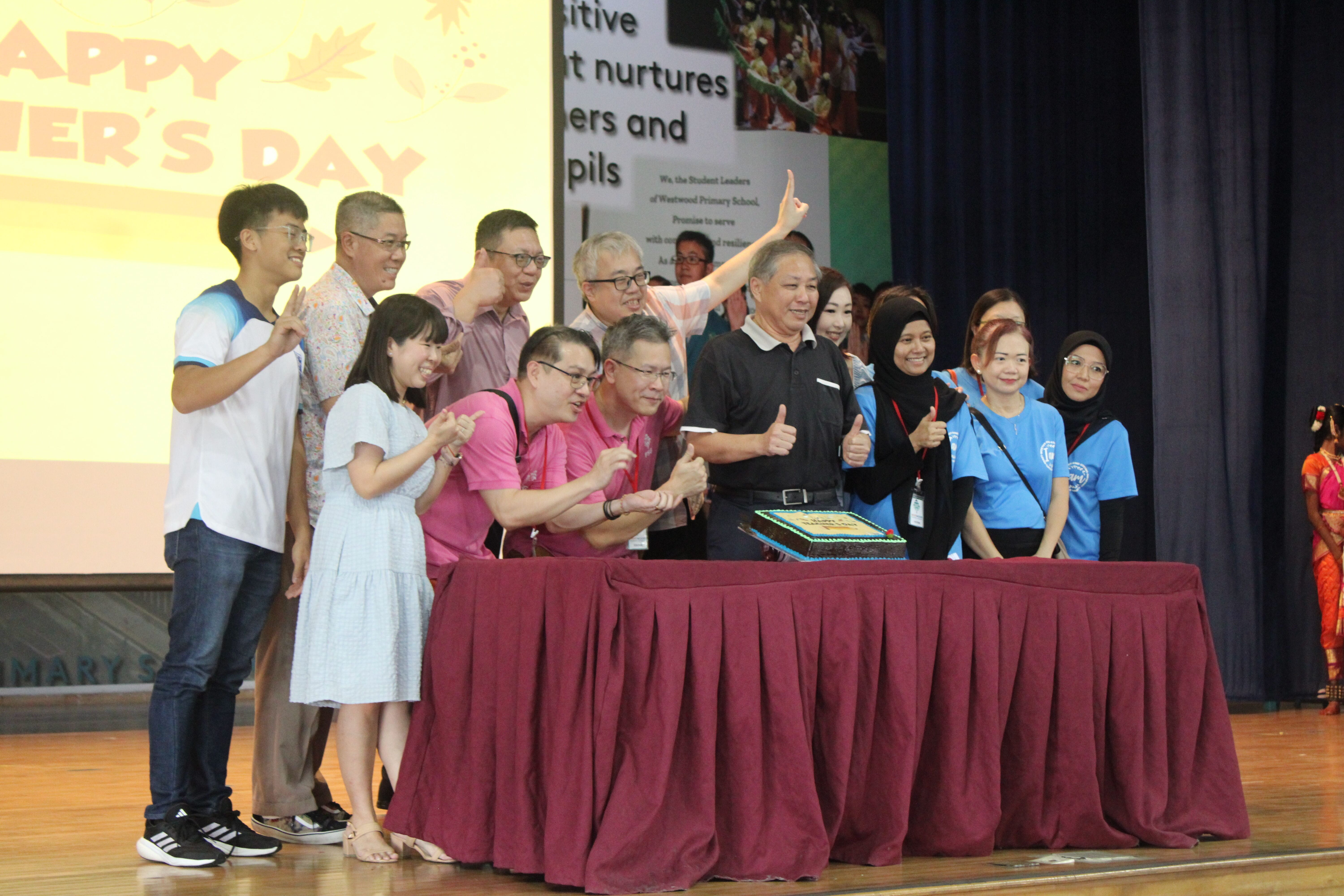 Group of people posing with thumbs up around a cake on a red-draped table at a Teachers Day celebration.
